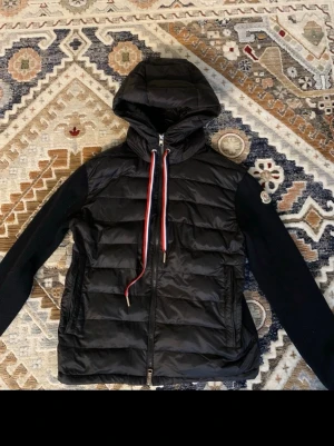 Svart Moncler cardiganjacka med huva - Svart cardiganjacka från Moncler med vadderad front och huva, stickade ärmar och rygg. Jackan har dragkedja framtill, två fickor och detaljer i rött och vitt på snörena. Klassisk Moncler-logga på ena ärmen. Perfekt för kyliga dagar. Använd max 5 gånger, säljer pga har blivit för liten. Pris kan gå ner vid snabbaffär. pm vid frågor