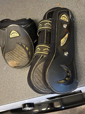 Svarta benskydd till häst Veredus - Säljer ett par svarta benskydd till häst från Veredus Grand Slam med kolfiberdetaljer och kardborreband. Skydden har guldfärgade loggor och detaljer, samt slitstark plast och vadderad insida för extra komfort. Se bilderna för slitage. Storlek M både fram och bak. Kontakta mig gärna om du har frågor🥰🥰