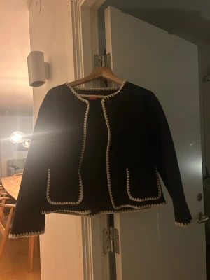 Svart cardiganjacka från RYVLS - Stilren svart cardiganjacka från RYVLS med dekorativa vita kanter och två stora fickor framtill. Jackan har rund halsringning och raka ärmar. Perfekt för dig som gillar en klassisk men trendig look. Nypris 600kr. 💕 Passar även xs