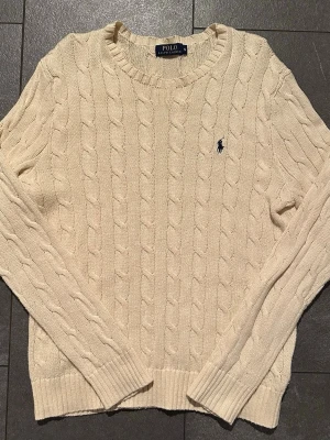 Kabelstickad offwhite tröja Polo Ralph Lauren - Kabelstickad offwhite tröja från Polo Ralph Lauren med rund halsringning och klassisk blå logga broderad på bröstet. Tröjan har långa ärmar och ribbade muddar vid ärmslut och nederkant. Perfekt för en stilren och avslappnad look.