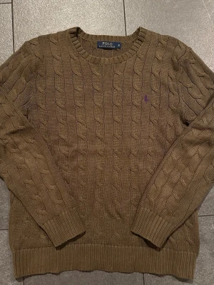 Brun kabelstickad tröja Polo Ralph Lauren - Kabelstickad tröja i brun nyans från Polo Ralph Lauren. Klassisk rund halsringning, ribbade muddar och lila broderad logga på bröstet. Tillverkad i mjukt material som passar perfekt till höst och vinter.