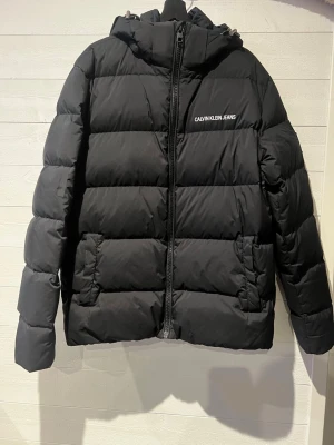 Svart pufferjacka Calvin Klein Jeans - Svart pufferjacka från Calvin Klein Jeans med huva och dragkedja framtill. Jackan har logotyp på bröstet och ärmarna, två stora fickor fram och quiltad design. Perfekt för kalla vinterdagar och riktigt snygg till streetwear-stilen. Den är lagad på ena armen