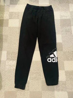 Svarta mjukisbyxor från Adidas - Svarta mjukisbyxor från Adidas med vit logga på ena benet. Byxorna har resår i midjan och vid bensluten samt två fickor fram. Perfekta för en avslappnad och sportig stil.