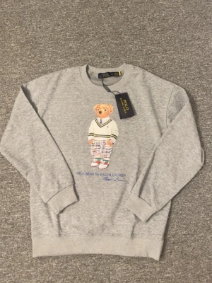 Grå sweatshirt Polo Bear Ralph Lauren - Grå sweatshirt från Ralph Lauren med ikoniska Polo Bear-trycket framtill. Rund halsringning, ribbade muddar och avslut. Tillverkad i mjuk bomullsblandning. Perfekt för en avslappnad och trendig look.