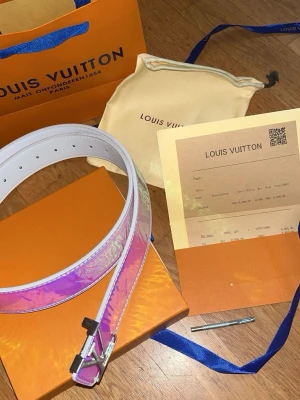 Louis Vuitton holografiskt bälte - Säljer ett Louis Vuitton bälte med holografisk yta i lila och rosa toner. Bältet har silverfärgat spänne och vita kanter. Kommer med originalpåse, box och kvitto. Perfekt accessoar för att lyfta din outfit.