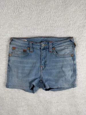 True Religion Jennie Curry Jeansshorts 30 Mid Rise Skinny - Säljer ett par ljusblå jeansshorts med klassisk femficksdesign och snygga kontrastsömmar. Shortsen har knappgylf och bälteshällor, samt dekorativa knappar på fickorna. Perfekta för varma dagar och enkel att matcha med olika stilar.