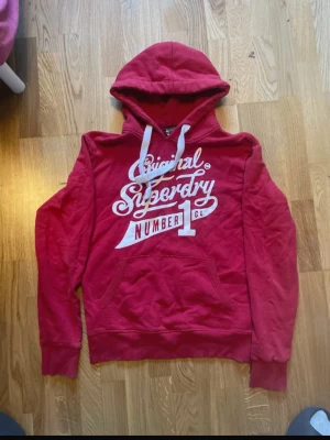 Röd hoodie från Superdry - Säljer en röd hoodie från Superdry med vit tryckt logga och text på bröstet. Hoodien har huva med vita snören och en stor magficka. Mjuk insida och ribbade muddar vid ärmar och nederkant. Perfekt för en avslappnad och cool stil.