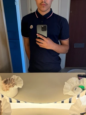 Mörkblå pikétröja med rödvit kant - Snygg mörkblå pikétröja med krage och knappar framtill. Tröjan har röda och vita detaljer på krage och ärmslut samt en broderad logga på bröstet. Perfekt för en stilren och sportig look.