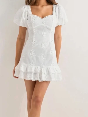 Vit kort klänning med volang - Söt vit kort klänning med puffärmar, volangkant nertill och broderade blommor. Klänningen har öppen rygg med stor rosett och fina spetsdetaljer längs kanterna.