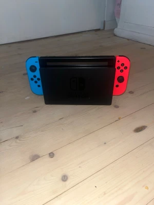 Nintendo Switch med blå och röd Joy-Con - Nintendo Switch spelkonsol med neonblå och neonröd Joy-Con-kontroller. Konsolen har en svart dockningsstation och är tillverkad i plast. Perfekt för gaming både hemma och på språng. Och massa spel ingår!