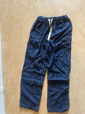 Mörkblå pyjamasbyxor med mönster - Säljer ett par mörkblå pyjamasbyxor med små vita mönster och vit dragsko i midjan. Byxorna har avslappnad passform och är perfekta för chill dagar hemma. Tillverkade i mjukt tyg för extra komfort.