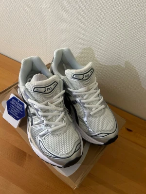 Vita Asics GEL sneakers med silverdetaljer - Säljer ett par helt nya ASICS GEL-KAYANO 14 i en stilren silver/vit färg. Skorna är oanvända och levereras i originalkartong.  GEL-KAYANO 14 är en populär unisexmodell som kombinerar en retroinspirerad design med modern komfort. Skon har en ventilerande vävd ovandel, detaljer i syntetiskt läder och en slitstark gummiyttersula som ger bra grepp. Den välkända GEL-dämpningen ger hög komfort och effektiv stötdämpning, vilket gör skorna perfekta för både vardagsbruk, promenader och längre dagar.