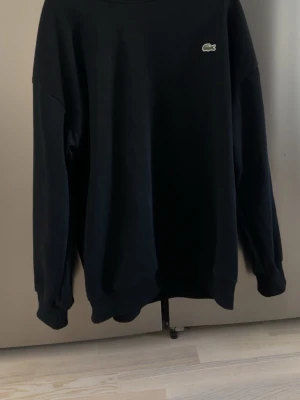 Svart sweatshirt från Lacoste - En stilren svart sweatshirt från Lacoste med rund halsringning och diskret krokodillogga på bröstet. Tröjan har långa ärmar och ribbade muddar vid ärmslut och nederkant. Perfekt för en avslappnad och trendig look.