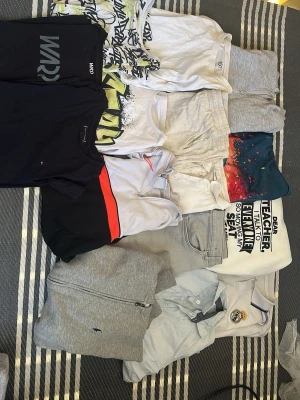 Mix av t-shirts, tröjor och byxor - Säljer ett paket med flera plagg: t-shirts i vitt, svart och färgglada tryck, en ljusgrå ziptröja, ljusgråa byxor, samt en vit piké med Real Madrid-logga. Plaggen har olika tryck och detaljer, vissa med sportiga inslag och grafiska motiv. Perfekt för dig som gillar en avslappnad och sportig stil. Med storlekar från 146-170