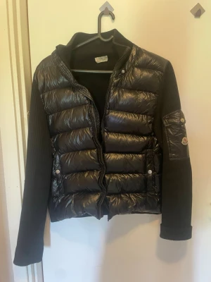 Svart pufferjacka med stickade ärmar - Snygg svart pufferjacka med glansig vadderad front och detaljer på axlar och ärm. Ärmarna är stickade och har en patch på vänster arm. Jackan har dragkedja framtill och två fickor med knappar.