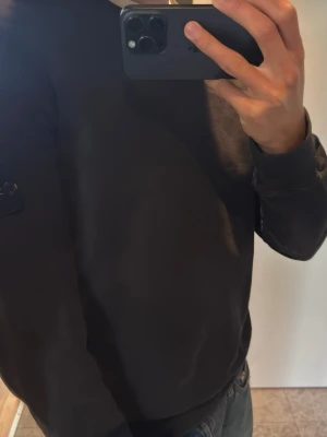 Svart sweatshirt från Stone Island - Svart sweatshirt från Stone Island med klassisk rund hals och ribbade muddar. Tröjan har den ikoniska logotypen på vänster ärm och är tillverkad i mjukt bomullsmaterial. Perfekt för en avslappnad och stilren look.