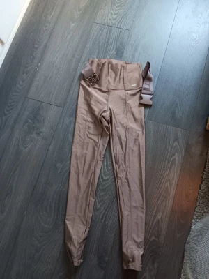 Bruna tights med hög midja och bälte - Säljer ett par bruna tights med hög midja och ett brett elastiskt bälte med spänne. Tightsen har en figurnära passform och är tillverkade i ett stretchigt material som passar perfekt för träning eller vardagsaktiviteter. Snygg detalj med sömmar framtill. Defekt med en söm som bild visar 
