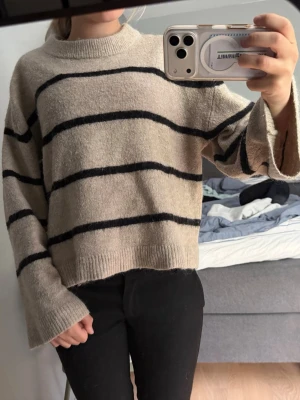 Beige stickad tröja med svarta ränder - Säljer en beige stickad tröja med breda svarta horisontella ränder. Tröjan har en avslappnad passform, långa ärmar och ribbade muddar vid hals, ärmslut och nederkant. Perfekt för en chill och stilren look.