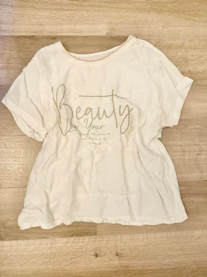 Vit t-shirt med textprint M/L - Vit t-shirt med korta ärmar och rund hals. Framsidan har ett stilrent texttryck i silver med orden 'Beauty Be Your'. Tillverkad i mjuk bomull, perfekt för en avslappnad look. Etiketten är borta men storleken ska vara M/L