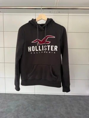Svart hoodie från Hollister - Svart hoodie från Hollister med stor vit logga och röd fågel på bröstet. Klassisk känguruficka framtill och justerbar huva med dragsko. Mjuk bomullskänsla och ribbade muddar vid ärmslut och nederkant. Perfekt för en avslappnad stil.