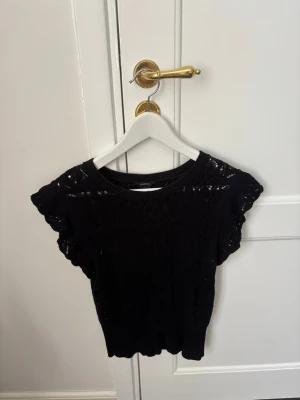 Lindex - Svart topp från Lindex med korta ärmar och vackert spetsstickat mönster. Toppen har rund halsringning och är perfekt för dig som gillar en feminin och stilren look. Materialet känns mjukt och luftigt, vilket gör den skön att bära.
