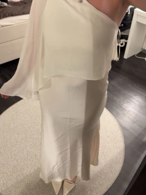 Vit oneshoulder långklänning - Elegant vit långklänning i oneshoulder-modell med svepande tygdetalj över axeln. Klänningen har en slät, figurnära passform och är tillverkad i ett glansigt material som ger en lyxig känsla. Perfekt för speciella tillfällen. Använd enbart två gånger och tvätt innan den skickas 