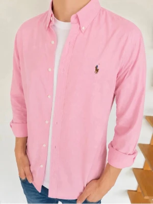 Rosa skjorta från Ralph Lauren - Klassisk rosa skjorta från Ralph Lauren med button-down krage och broderad logga på bröstet. Skjortan har långa ärmar, vita knappar och är tillverkad i bomull. Perfekt för en clean och stilren look.