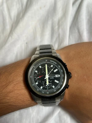 Seiko kronografklocka i silver - Stilren Seiko kronografklocka med svart urtavla, silverfärgat armband i metall och tydliga vita visare. Klockan har datumvisning och flera små urtavlor för extra funktioner. Perfekt accessoar för dig som gillar klassisk och sportig stil. Jag fick den av min pappa då han har använt den lite, förut när han hade den, hittade den sittandes några dagar sen och den fungerar inte, jag vet inte vad felet är men det går nog att fixa då den har funkat innan!