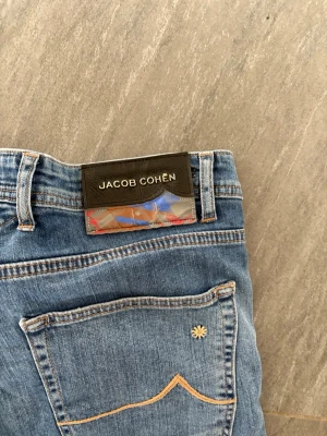 Blå Jacob Cohën jeans - Snygga blå jeans från Jacob Cohën med raka ben. Jeansen har ljusblå tvätt, kontrastsömmar och en unik detalj med liten blomma på bakfickan. Tillverkade i mjukt denimtyg för en bekväm känsla. Pris går att diskutera vid snabb affär 😊