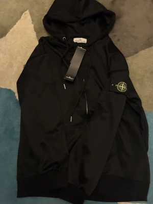 Svart hoodie från Stone Island - Snygg svart hoodie från Stone Island med klassisk logga på ärmen. Hoodien har huva med dragsko och ribbade muddar. Tillverkad i mjukt material som känns skönt mot huden. Perfekt för dig som vill ha en stilren och trendig look.
