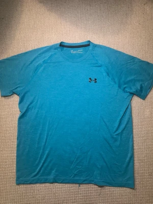 Blå t-shirt från Under Armour - Snygg blå t-shirt från Under Armour med klassisk logga på bröstet. T-shirten har rund halsringning och korta ärmar. Perfekt för träning eller chill dagar. Tillverkad i ett lätt och mjukt material som andas.