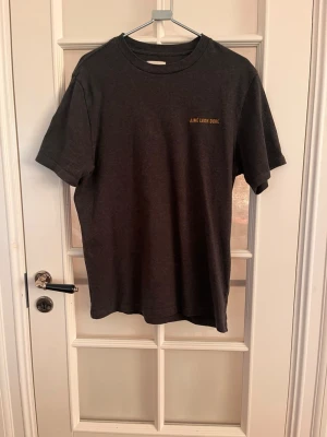 Mörkgrå t-shirt från Aimé Leon Dore - Snygg mörkgrå t-shirt från Aimé Leon Dore med diskret gul text på bröstet. Klassisk passform med rund hals och korta ärmar. Perfekt basplagg för en clean och stilren look.