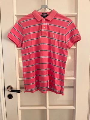 Rosa randig pikétröja från Polo Ralph Lauren - Säljer en rosa pikétröja med vita och blå horisontella ränder från Polo Ralph Lauren. Klassisk krage med två knappar, korta ärmar och broderad logga på bröstet. Tillverkad i mjuk bomull för en bekväm känsla.