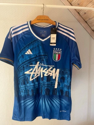Italien fotbollströja Adidas x Stüssy - Snygg blå fotbollströja från Adidas i samarbete med Stüssy och Italien. Tröjan har vita och guldiga detaljer, Italiens landslagsemblem, samt ett stort tryck av Colosseum och Stüssy-loggan på framsidan. Tillverkad i lätt och ventilerande material.