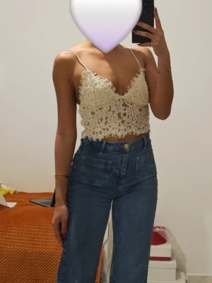 Beige spetsig croppad linnetopp - Säljer en beige croppad linnetopp med tunna axelband och v-ringning. Toppen är täckt av vacker spets och har en feminin, bohemisk vibe. Perfekt för sommardagar eller festivaler. Passar dig som gillar detaljer och en lätt, luftig känsla.