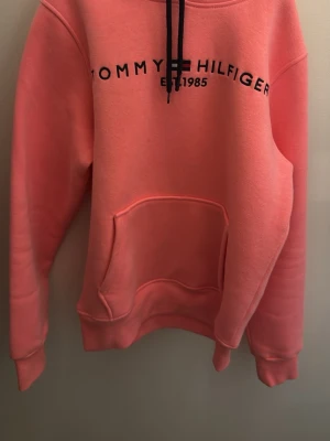 Rosa hoodie från Tommy Hilfiger - Säljer en snygg rosa hoodie från Tommy Hilfiger med svart logga och text på bröstet. Hoodien har huva med svarta snören och en stor magficka framtill. Perfekt för en chill och avslappnad stil. Det är storlek l men passar mig som har xs/s.