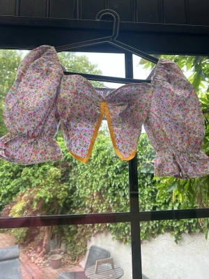 Blommig puffärm topp med volang - Supersöt blommig blus med stora puffärmar och volangdetaljer. Toppen har en öppen front med gul kant och elastisk rygg för skön passform. Perfekt för dig som vill sticka ut med en färgglad och lekfull look i sommar.