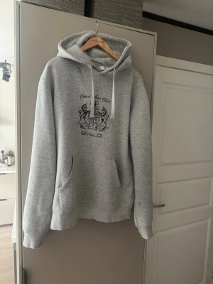 Grå hoodie med tryck från BVALDI - Snygg grå hoodie från BVALDI med svart tryck framtill som visar ett vapensköld-motiv och texten 'Côte d'Azur Club'. Hoodien har huva med dragsko och en stor magficka. Perfekt för en chill och avslappnad stil.