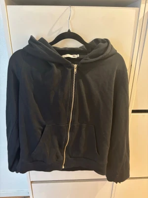 Svart hoodie med dragkedja och huva - En enkel svart hoodie med dragkedja framtill och stor huva. Hoodien har två rymliga fickor på magen och är tillverkad i mjukt sweatshirtmaterial. Perfekt för en avslappnad stil och funkar året runt.