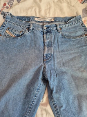 Vintage jeans från Diesel - Säljer ett par klassiska ljusblå jeans från Diesel med raka ben och fem fickor. Jeansen har snygga kopparfärgade nitar och en liten Diesel Industry-logga på myntfickan. Perfekta för en avslappnad och trendig look.