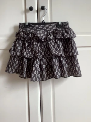 Svart volangkjol med mönster - Kort svart kjol med vita mönster och volanger. Kjolen har resår i midjan och inbyggda shorts. Den är endast använd ett fåtal gånger så nästan nyskick