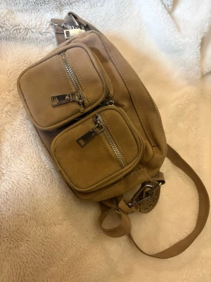 Beige shoppingväska med nitar - Beige shoppingväska i mockaimitation med två framfickor och silvriga dragkedjor. Axelremmen är dekorerad med nitar för en trendig look. Perfekt för dig som gillar praktiska detaljer och stilren design.