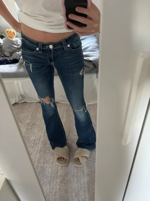 Blå bootcut jeans med slitningar - Säljer ett par blå bootcut jeans med coola slitningar och hål på benen. Jeansen har låg midja, klassiska fickor och vita kontrastsömmar. Perfekta för dig som gillar en avslappnad och trendig stil.