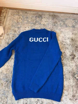 Blå stickad tröja  - Blå stickad tröja från Gucci med vit logga på ryggen. Tröjan har rund halsringning, långa ärmar och ribbstickade muddar vid ärmslut och nederkant. Perfekt för dig som vill ha en statement piece i garderoben. Stor small på den men passar mer som M/L