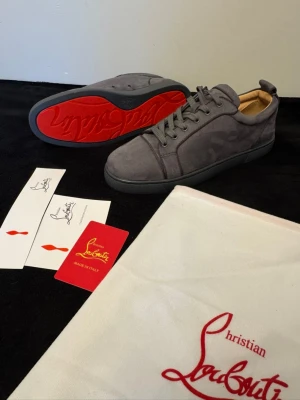 Gråa sneakers från Christian Louboutin - Snygga gråa sneakers i mocka från Christian Louboutin med den ikoniska röda sulan. Skorna har lågt skaft, ton-i-ton snörning och diskreta sömdetaljer. Perfekta för dig som vill ha en exklusiv och stilren look.