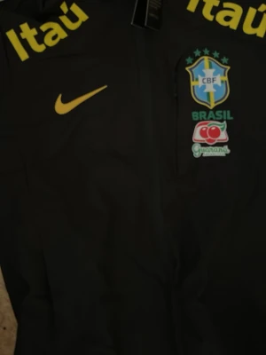 Svart Brasil Nike vindjacka med huva - Svart vindjacka från Nike med huva och dragkedja. Jackan har Brasil-logga, gula och röda tryck som 'Itaú' och 'vivo' samt GORE-TEX-material för extra väderskydd. Perfekt för dig som gillar sportig stil och vill sticka ut med landslagskänsla.