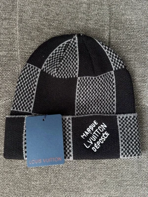 Svart och grå mössa - Säljer en snygg svart och grå mössa med rutigt mönster och broderad text på uppviket. Mössan är stickad och har en modern, trendig look som passar perfekt till alla säsonger.