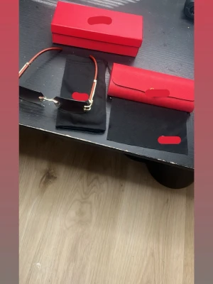 Guldiga solglasögon med röda bågar - Säljer ett par stilrena solglasögon med guldiga detaljer och röda bågar. Glasen är rektangulära och har en lätt tonad röd färg. Kommer med rött fodral och svart putsduk. Perfekta för dig som vill sticka ut med en trendig accessoar.