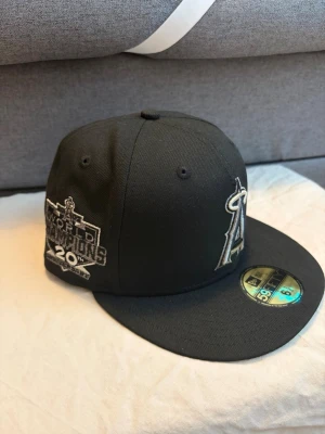 Svart New Era LA Angels keps - Säljer en svart keps från New Era med LA Angels-logga broderad framtill i vitt och silver. Kepsen är en 59FIFTY-modell med platt skärm och sticker kvar på ovansidan. På sidan finns broderad text om World Champions. Helt ny säljer för att passar ej varan är köpt 26/03/29