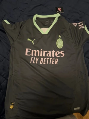 AC Milan matchtröja  - Snygg AC Milan matchtröja från Puma i mörkgrönt med ljusgröna detaljer. Tröjan har klubbmärke och Puma-logga på bröstet samt Emirates Fly Better-tryck framtill. Tillverkad i lätt och ventilerande material, perfekt för fotboll eller träning.